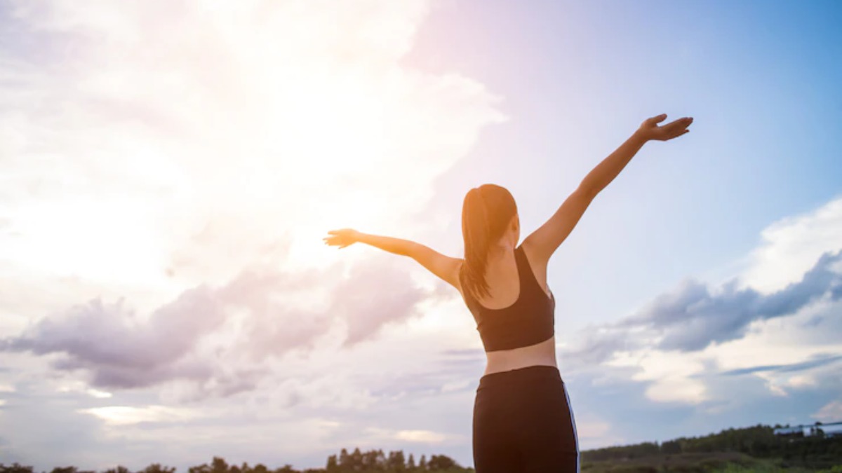 Woman in athletic wear seen from behind with arms raised toward a bright cloudy sky, silhouette style suggesting civic energy and community support