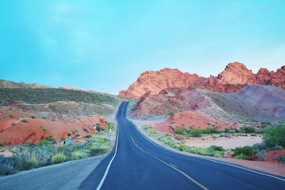 A paved road curving through a wide open landscape with rock formations under a blue sky, evoking a sense of a long district campaign trail