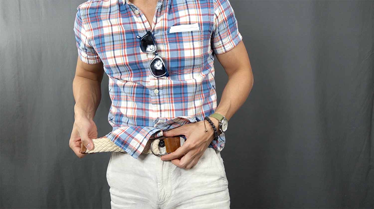 Torso of a man in a plaid short-sleeve shirt adjusting a woven belt over white pants against a gray curtain background, suggesting an everyday working candidate